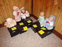 Five Knowles Yolanda dolls seated on black boxes with name tags, displayed on carpet against wood panel wall.