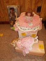 Photo showing all three dolls: Anne of Green Gables doll in box, Christmas Angel pillow doll seated in pink fabric chair, Knowles Megan doll lying down.