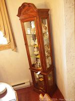 Full view of wood and glass curio cabinet in corner beside a window, showing front and part of one side with displayed figurines inside.