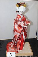 Full view of Japanese Geisha doll in red kimono and white headpiece on wooden stand.