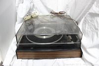 Closed record player showing wood grain base and clear smoked plastic dust cover with attached power cords on top.