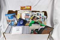 Wide view showing large cardboard box filled with assorted trading cards, loose and sleeved, including baseball and other cards. Includes unopened Topps baseball card packs.