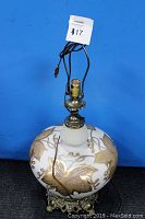 Photo of vintage table lamp showing rounded white glass base with gold leaf etching, ornate brass base, and electrical cord attached.