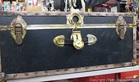 Front view of vintage foot locker showing central brass lock plate, latch, and black exterior with copper riveted trim.