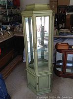 Front and side view showing green wooden cabinet with glass sides and two upper glass shelves. Lower part has wooden door cupboard. Cabinet is tall and slim with decorative beaded trim on the glass frames.