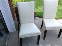Front and side view of two white leather Parsons chairs with dark wood legs pictured outside on concrete