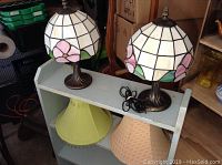 Two Tiffany style lamps and two fabric shades on a shelf. Lamps are stained glass with floral pattern, shades are yellow-green and beige.