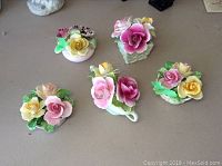 Five porcelain floral bouquets arranged on a table showing varied designs and pastel colored roses with green leaves.