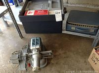 Viewlex slide projector on the floor near a large plastic carrying case and a blue suitcase, showing the metallic grey body and slide holders on projector.