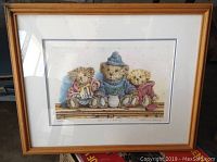 Framed print of three teddy bears sitting on a wooden bench, signed and numbered, with visible matt and frame.