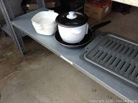 Overview of the Rival slow cooker with black lid and base, two white Corning Ware dishes, and a black non-stick skillet on a metal shelf.
