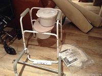 Photo shows adjustable metal commode chair with armrests and removable bucket, spare bucket with lid, and new toilet seat in packaging on floor near furniture.