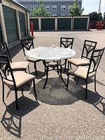 Outdoor photo showing round glass kitchen table with black iron pedestal and matching 6 chairs with beige cushions.
