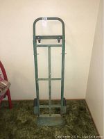 Front view of the green metal hand truck with handle, frame, and wheels on a carpeted floor.