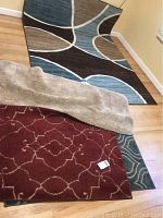 Photo showing three different rugs: blue and brown abstract patterned rug, red patterned rug partially covered by a beige shaggy rug, and a corner of a greenish rug on hardwood floor