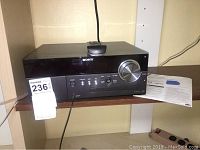 Photo of the Sony micro hi-fi main unit on shelf showing front panel with volume knob and input buttons, beside user manual.