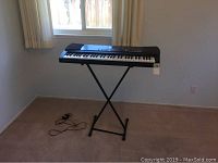 Full view of black CASIO CT 555L keyboard on X-style stand in room with window and curtains, power adapter on floor.