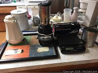 Photo showing the full lot on a counter featuring blender, carving knife box, scale, serving tray with whisk illustration, and other kitchen items