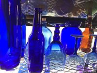 Blue glass bottles, stemware including wine glasses, vases, and a brown Grolsch beer bottle on a wire shelf with sunlight behind them.