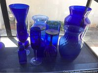 Photo showing a group of nine blue glass items including vases, stemware glasses, and a small bottle arranged on a surface against a window.