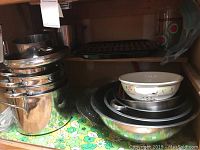Stack of stainless steel pots with lids along with nested metal mixing bowls and ceramic bowl on top, visible floral pattern on small bowl, all placed on a shelf with floral shelf liner.