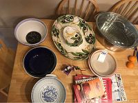 Photo showing multiple ceramic bowls, a large ceramic serving platter with olive branch design, and a metal roasting pan with lid on a wooden table.
