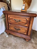 Wooden night stand with two drawers and metal handles, placed on carpet near bed and power strip, showing overall shape and condition
