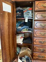 Open wooden wardrobe showing neatly folded men's clothing including T-shirts and jeans in neutral colors.