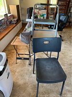 Photo showing black plastic chair, part of the camp roll table and folding table behind it, also silver step ladder on top of the camp table.