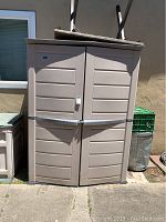 Front view of beige plastic outdoor tool shed with two doors, latch mechanism, and weathering marks.