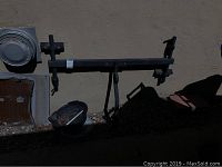 Photo of a black metal car bike rack with adjustable clamps against an exterior beige wall, showing the entire structure lying flat on the ground with some outdoor items in the background.