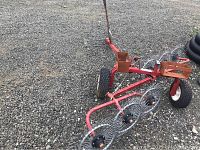 Full view of red metal hay tedder rake mounted on wheels with hitch on gravel surface.