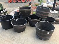 Photo showing all seven planters arranged outdoors on concrete ground, including several large plastic wine barrel style pots and smaller round pots.