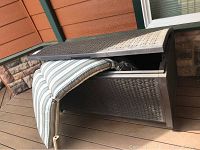 Brown Suncast outdoor storage chest with hinged lid partially open, woven wicker pattern on the sides, placed on a deck near an orange wall.
