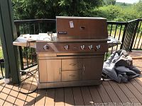 Full view of stainless steel grill on deck, side shelves folded out, cover and accessories on the side