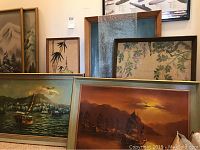 Six framed artworks displayed together on floor and leaning against wall showing variety of subjects and frame styles.
