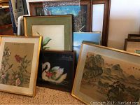 Group view of several framed paintings and prints leaning against a wall, showing floral and Asian-style art.