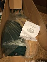 Boxed new hanging hammock chair components including green fabric seat, cushions, wooden bar wrapped in bubble wrap, and assembly instructions.