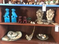 Overview of shelf with blue and reddish foo dog pairs, two soapstone floral carvings, small elephant and koi, plates, glass eggs, and bird figurines