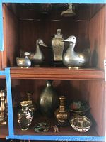 Photo showing pewter duck dishes, pewter lidded urn, and cloisonne urn and decor pieces in a wooden cabinet.