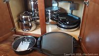Opened cabinet with stacked kitchenware: stainless steel pots, T-Fal fry pans, glass lids, and a cast iron griddle on the bottom shelf.