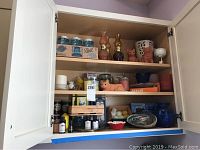 Full cabinet view showing all items including boxed Ball jars on top shelf, oil lamps, cat figurines, candles and abalone shell on bottom shelf.