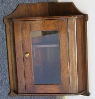 Closed view of the antique corner shelf showing carved wood, glass door with brass knob, and shaped top edge.