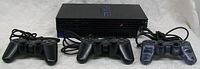 Front view showing PlayStation 2 console with three wired controllers in front: two black and one translucent gray.