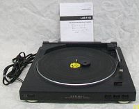 Optimus LAB-1100 fully automatic belt drive turntable shown front view with instruction manual behind unit. Power cord visible. Black base with integrated black platter and tonearm, yellow 45 RPM adapter on spindle.