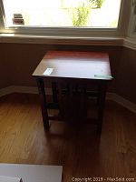 Front view of solid wood end table by window showing flat top surface and lower shelf, displaying wear and scratches.