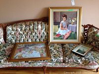 Three framed artworks on floral upholstered couch: two paintings and one print. Largest painting (girl with doll) centrally placed upright. Other painting (table setting) and print lying flat.