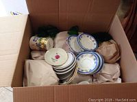 Open box showing stacked saucers, plates, and cups with floral patterns along with green glassware wrapped in protective paper.