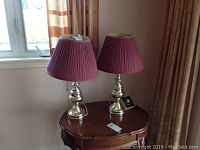 Two table lamps with silver-tone bases and purple pleated fabric shades placed on a wooden side table near a window.