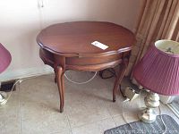 Image shows the full oval wooden end table with four curved legs and medium brown finish in room setting.